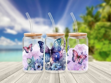 Drei 16 oz Glasbecher mit Bambusdeckel und Glastrinkhalm, dekoriert mit farbenfrohen Schmetterlings- und Blumenmotiven in Blau-, Lila- und Rosatönen. Die Becher stehen auf einem Holztisch vor einem Hintergrund mit Strand und Palmen, was ein Gefühl von Som
